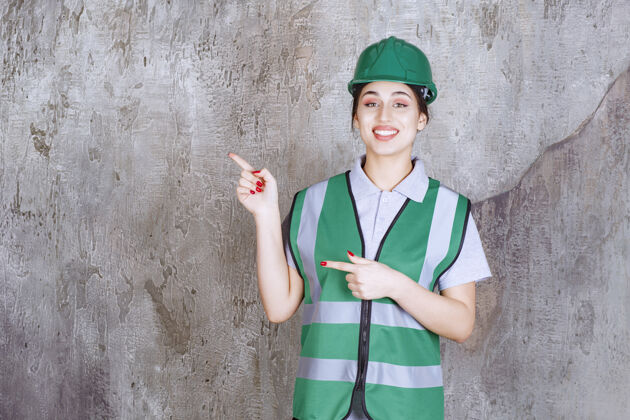 聪明身着绿色制服 头戴头盔的女工程师 表情丰富地展示着左侧方向年轻维修