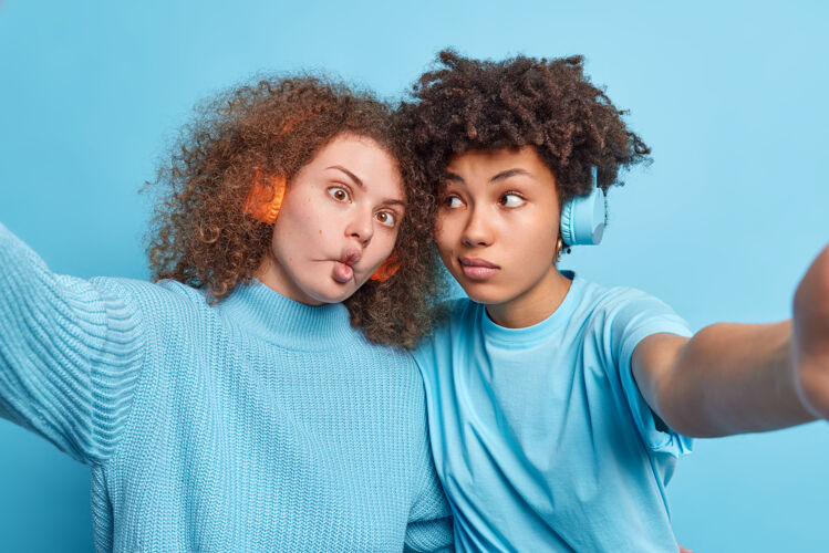 青少年有趣的不同女性的水平镜头做有趣的面部姿势自拍通过耳机听音乐站在蓝色墙壁旁边最好的混血朋友在室内玩得开心夫妇毛衣姿势