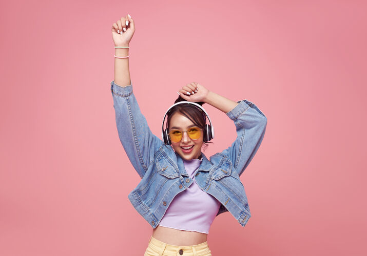 音乐快乐美丽的亚洲少女戴着黄色眼镜 戴着耳机听音乐 在粉红色的墙上跳舞室内配件快乐