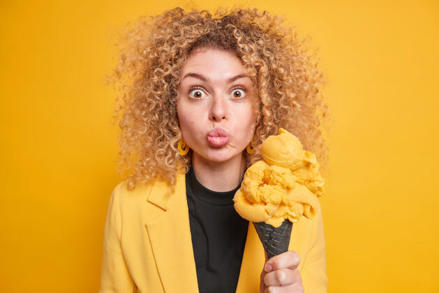 时尚惊讶的女人卷发保持嘴唇圆润举行开胃冰淇淋吃冷冷冻甜点在炎热的天气穿着时尚夹克隔离在黄色的墙壁夏天和垃圾食品食物女人室内