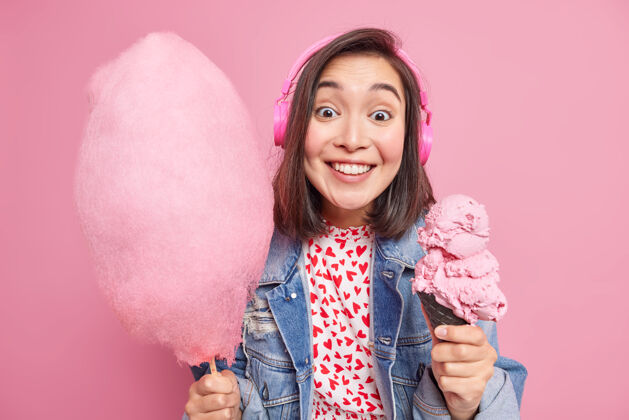 乐观漂亮 积极的深色头发的亚洲女模特微笑着 穿着时髦的衣服 在粉红色的墙上摆着美味可口的甜点享受冰淇淋和棉花糖吧快乐夏天反应