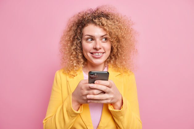 检查一位穿着公司服装 留着卷发 心事重重的女士手里拿着手机查看留言框 在社交媒体应用程序上发布在线聊天帖子 对着粉色的墙壁摆姿势通话模特短信