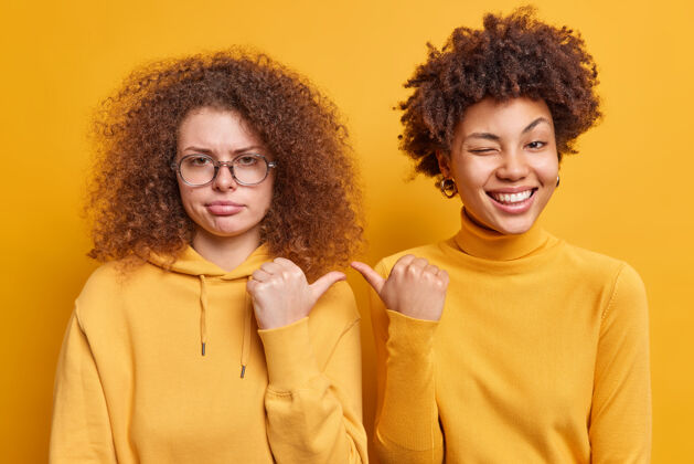 无忧无虑快乐的美国黑人妇女和她悲伤的卷发妹妹在黄色的墙壁上随意地穿上隔离的衣服 互相用拇指指着对方 表达不同的情感是她两个女人在室内可爱朋友非洲