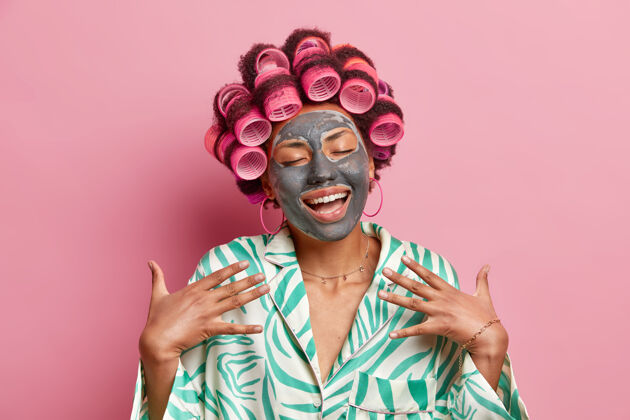 滚动满意的美国黑人女士笑容满面享受护肤治疗涂上滋养的粘土面膜让卷发发型穿上丝绸睡衣隔离在粉红色的墙上个人室内女人