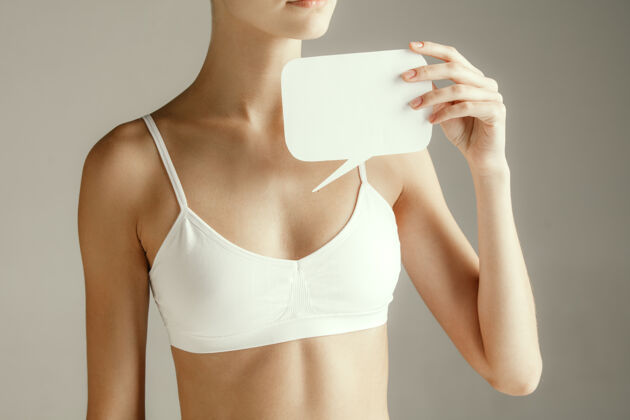 皮肤女性健康女模特拿着空卡片靠近乳房年轻的成年女孩用纸做标志或符号隔离在灰色工作室背景上切下身体的一部分医疗问题和解决方案胸围胸罩信息