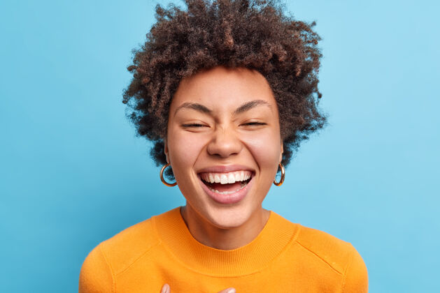 请美丽年轻的非洲裔美国女人的特写镜头 自然卷发 笑容灿烂 表情积极 心情愉快 穿着橙色的套头衫 隔着蓝色的墙非洲休闲非洲