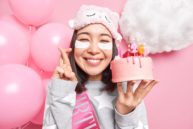 气球女人在生日蛋糕上吹蜡烛前许愿 手指交叉 微笑 穿着睡衣庆祝生日庆典室内眼罩