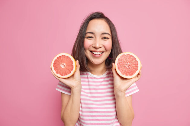 乐观可爱开朗的亚洲女人 深色头发 拿着两片葡萄柚 用柑橘类水果做天然化妆品 穿着随意 有快乐的表情 隔着粉色的墙壁素食人青少年