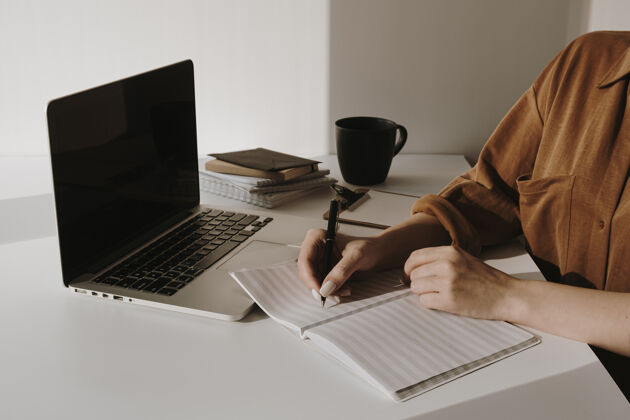 笔记女人在白纸上写字笔记本电脑桌子上有咖啡杯 纸床单 靠着白墙的文具业务手女人