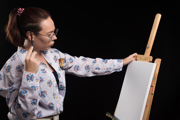画笔手握画笔和颜料的女艺术家站在黑色画架旁艺术家画笔手