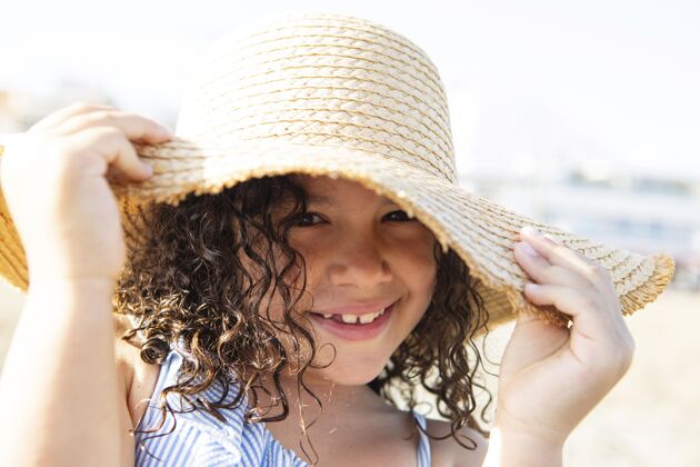 海边特写微笑女孩戴帽子在海滩上放松孩子户外