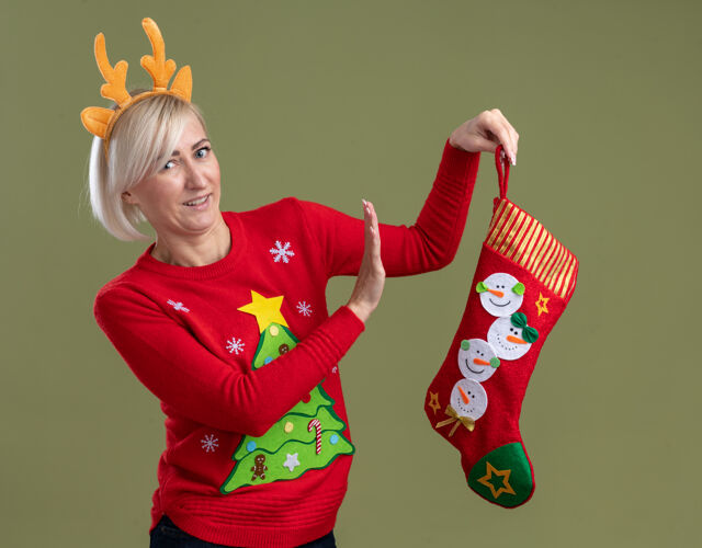 中年穿着圣诞驯鹿鹿角头带和圣诞毛衣拿着圣诞长袜的中年金发女人看起来像被隔离在橄榄绿的墙上驯鹿女人股票