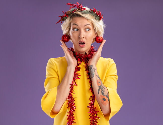 周围令人印象深刻的金发女郎戴着圣诞花环 脖子上戴着金箔花环 耳边挂着圣诞饰品 抚摸着紫色墙上孤立的饰品 还有复制空间装饰圣诞节穿