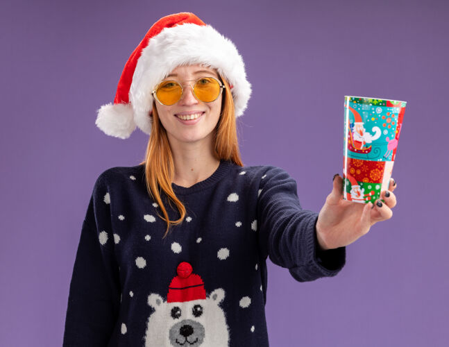 杯子微笑着的年轻漂亮的女孩穿着圣诞毛衣 戴着圣诞帽 戴着眼镜 把圣诞杯孤立在紫色的墙上帽子戴着拿着