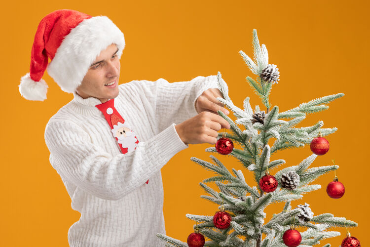 靠近高兴的年轻帅哥戴着圣诞帽 打着圣诞老人的领带站在圣诞树旁 用圣诞球装饰圣诞树 橙色的墙上孤立着请年轻人小伙子