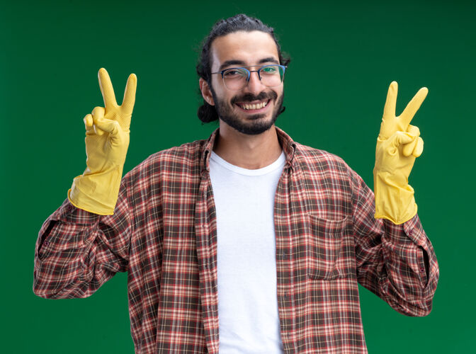 年轻微笑的年轻帅气的清洁工穿着t恤 戴着手套 在绿色的墙上显示出和平的姿态和平手势帅气