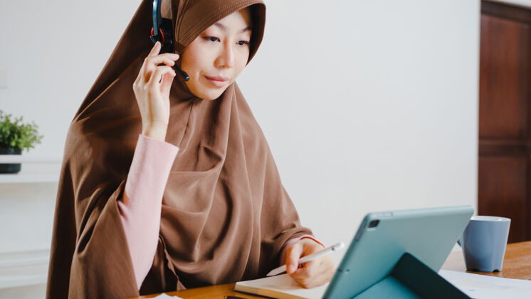 房子穆斯林女士戴着耳机使用数字平板电脑在家里厨房工作时 通过视频会议与同事谈论销售报告工作场所远程亚洲人