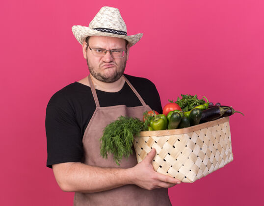 篮子皱着眉头的年轻男园丁戴着园艺帽 手里拿着蔬菜篮子 孤零零地站在粉红色的墙上感觉脸蔬菜