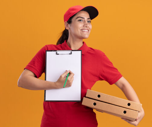 年轻人微笑着看着身边穿着制服 戴着帽子 拿着剪贴板 隔离在橙色墙上的比萨饼盒的年轻送货女孩衣服人拿着