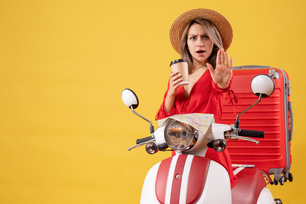 人正面图：身穿红色连衣裙的年轻女士拿着咖啡杯 在轻便摩托车旁示意停车标志漂亮游客手提箱