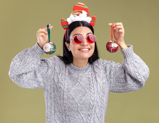 圣诞老人微笑着的年轻白人女孩戴着圣诞老人的头带 戴着一副眼镜 拿着圣诞饰品 靠近被隔离在橄榄绿墙上的头圣诞饰品头带穿