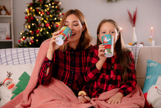 毯子高兴的母女俩抱着纸杯和饼干坐在铺着毯子的沙发上 在家里享受圣诞时光沙发抱着盖上