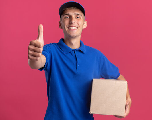 粉色微笑的年轻金发送货员竖起大拇指 拿着名片盒拇指向上拿着