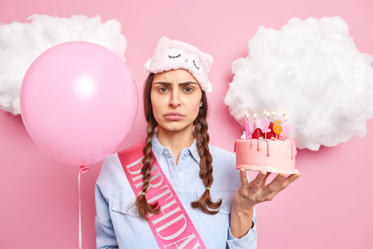 生日一个女人独自庆祝26岁生日 手里拿着草莓蛋糕和充气气球 穿着休闲的家居服 隔离在粉色上女性人沮丧