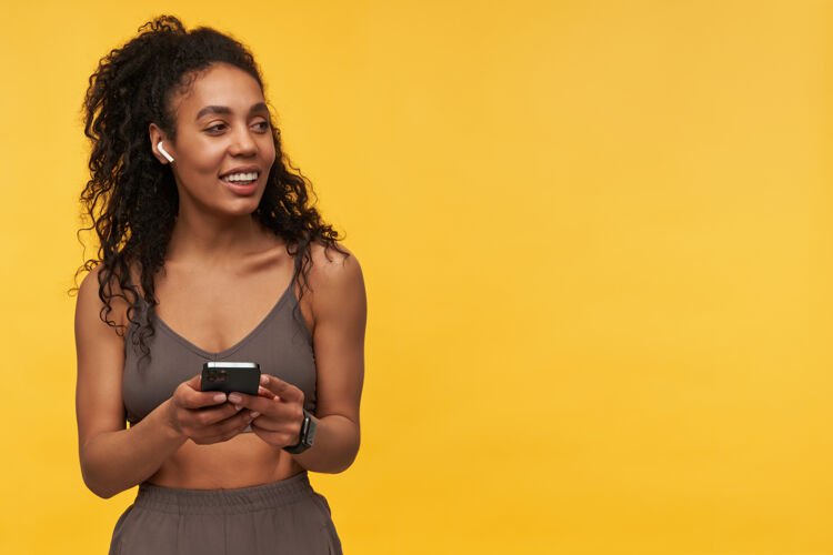 技术快乐美丽的年轻女运动员站在黄色的墙上 用无线耳机和手机听音乐女人版权空间卷发
