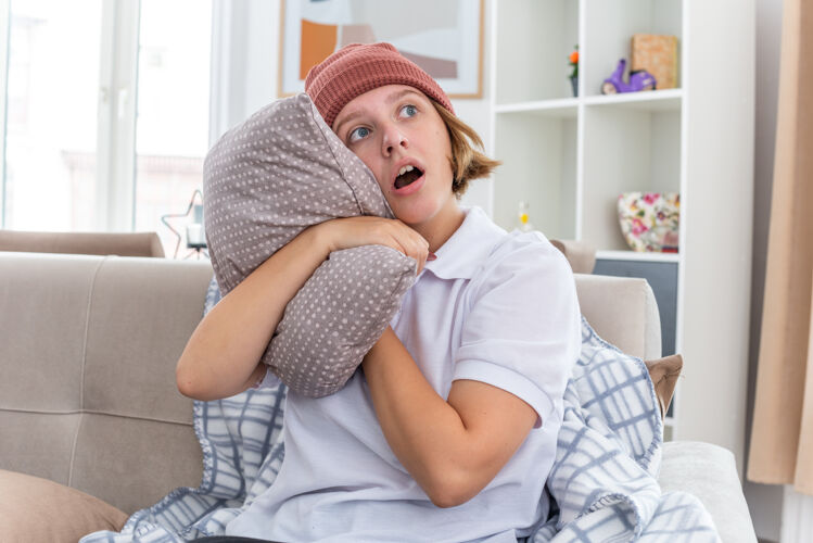 生活不健康的年轻女子戴着暖和的帽子 拿着毯子 看起来不舒服 生病 患感冒和流感 抱着枕头 坐在客厅里的沙发上忧心忡忡痛苦温暖旁边