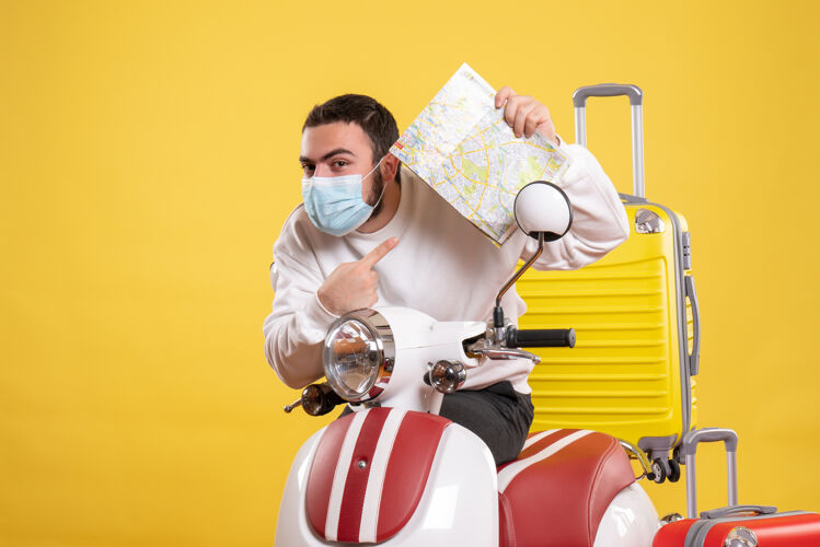 手提箱旅行概念的俯视图 戴着医用面罩的年轻人站在摩托车旁边 黄色手提箱放在上面 拿着地图感到好奇年轻人旅行感觉