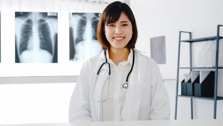 治疗自信的年轻亚洲女医生 身穿白色医疗制服 手持听诊器 看着摄像机 微笑着与健康医院的患者进行视频电话会议实验室看着摄像机医学