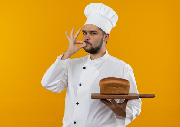 年轻自信的年轻男性厨师身着厨师制服 手持插有面包的砧板 在橙色的墙上做着与世隔绝的美味手势手握手势制服