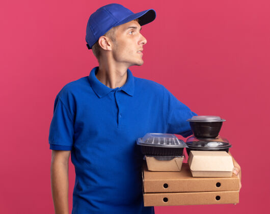 信心自信的年轻金发送货男孩拿着食物容器和比萨饼盒包装看一边食物比萨饼粉色