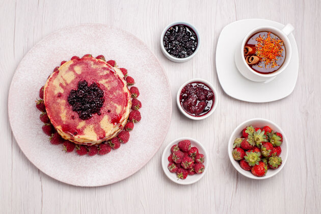 杯子俯瞰美味的草莓煎饼白桌上的一杯茶馅饼饼干甜浆果蛋糕派胡椒饼干