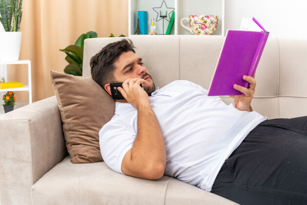 休闲一个穿着休闲服的年轻人抱着书 一脸严肃地读着书 讲着手机在家里度过周末 躺在客厅里的沙发上阅读年轻周末