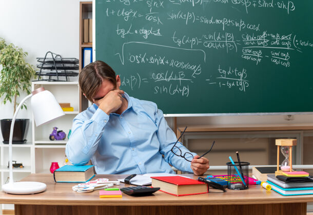 学校年轻的男老师戴着眼镜 劳累过度 用手捂着眼睛坐在课桌旁 在教室的黑板前放着书和笔记年轻劳累教室