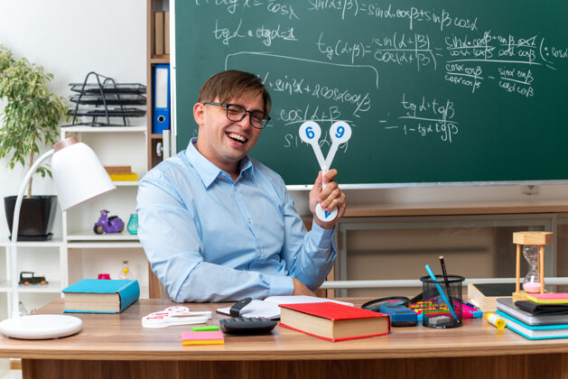 笔记年轻的男老师戴着眼镜 展示着解释课的车牌 微笑着坐在课桌旁 教室里的黑板前放着书和笔记黑板展示教室