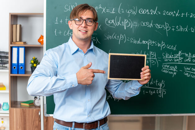 穿年轻的男老师戴着眼镜 拿着小黑板 用食指指着黑板 微笑着自信地站在教室里数学公式的黑板旁手指站指向