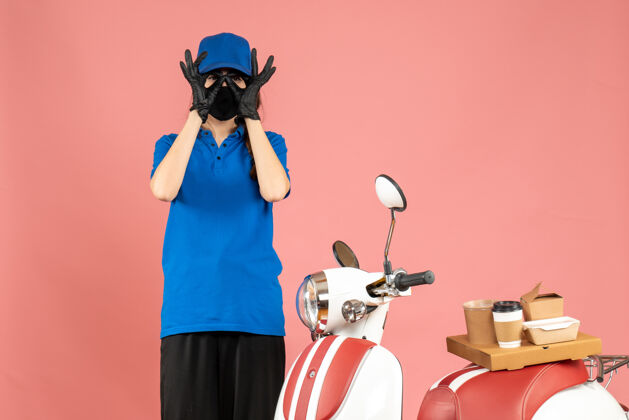 立场俯视图：戴着医用口罩的女快递员站在摩托车旁边 摩托车上放着咖啡蛋糕 在柔和的桃色背景上做着眼镜手势桃咖啡人
