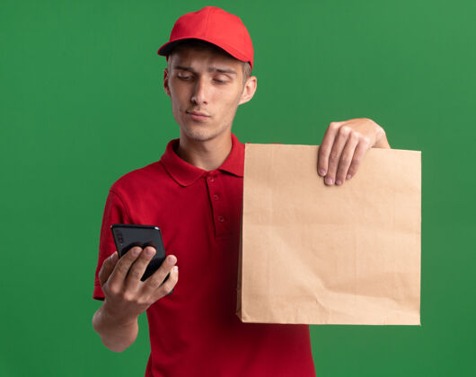 包装自信的年轻金发送货员拿着纸包看着电话电话纸持有