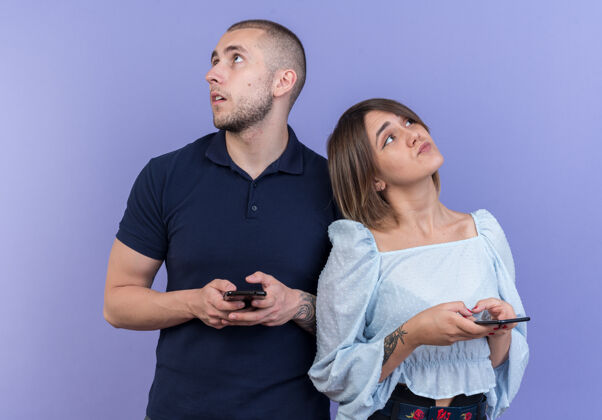 站立一对年轻漂亮的夫妇 带着智能手机 带着梦幻般的神情 站在蓝色的墙上 思考着积极的一面男人女人年轻