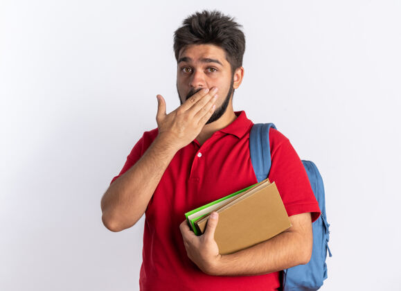 站立年轻的留着胡子的学生 穿着红色马球衫 背着背包 拿着笔记本 手捂着嘴站在白色的墙上封面年轻人个人