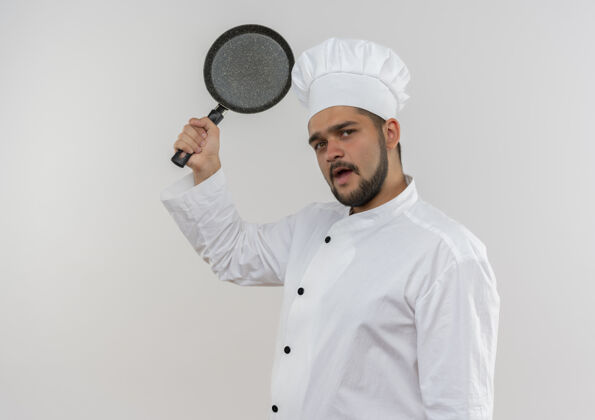 烹饪令人印象深刻的年轻男厨师在厨师制服提高煎锅隔离在白色墙壁与复制空间男薯条制服