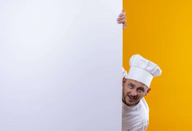 厨师穿着厨师制服的年轻帅哥站在白色的墙后 指着隔离在橙色墙上的复制空间帅气指点墙壁