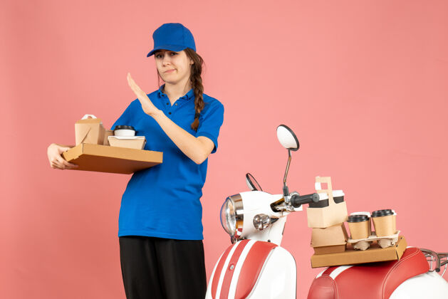 咖啡疲惫的快递员女孩站在摩托车旁 手里拿着咖啡和粉彩桃色背景上的小蛋糕举行背景粉彩