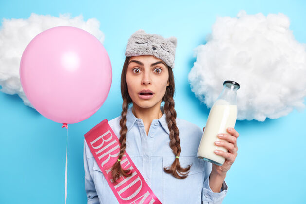 惊喜一个留着两条辫子的女人手里拿着粉红色的充气气球 一瓶装牛奶的玻璃瓶上戴着一条缎带 上面写着生日 蓝色的惊呆表情举行