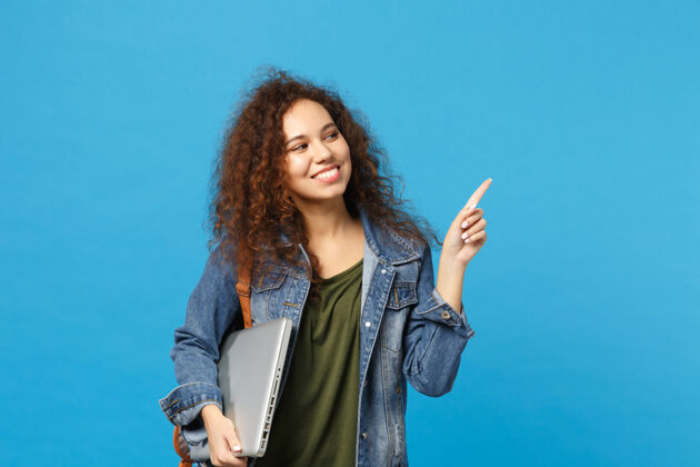 使用年轻的非洲裔美国女孩十几岁的学生在牛仔服装 背包举行电脑隔离在蓝色的墙壁上手势手指拿着