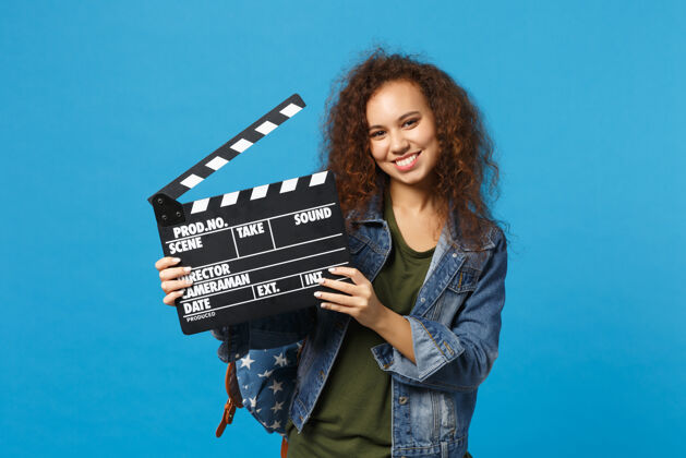 媒体年轻的非洲裔美国女孩十几岁的学生在牛仔服装 背包举行克拉珀隔离在蓝色的墙壁上人摄影剪辑