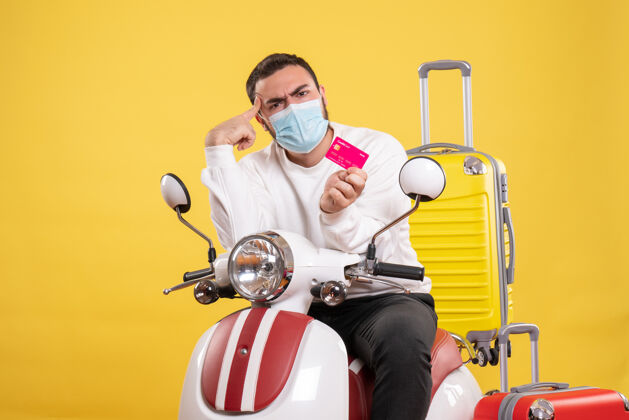 持有旅行概念的正面图 戴着医用面罩的年轻困惑的家伙坐在摩托车上 黄色手提箱在上面 手里拿着银行卡男性坐着黄色